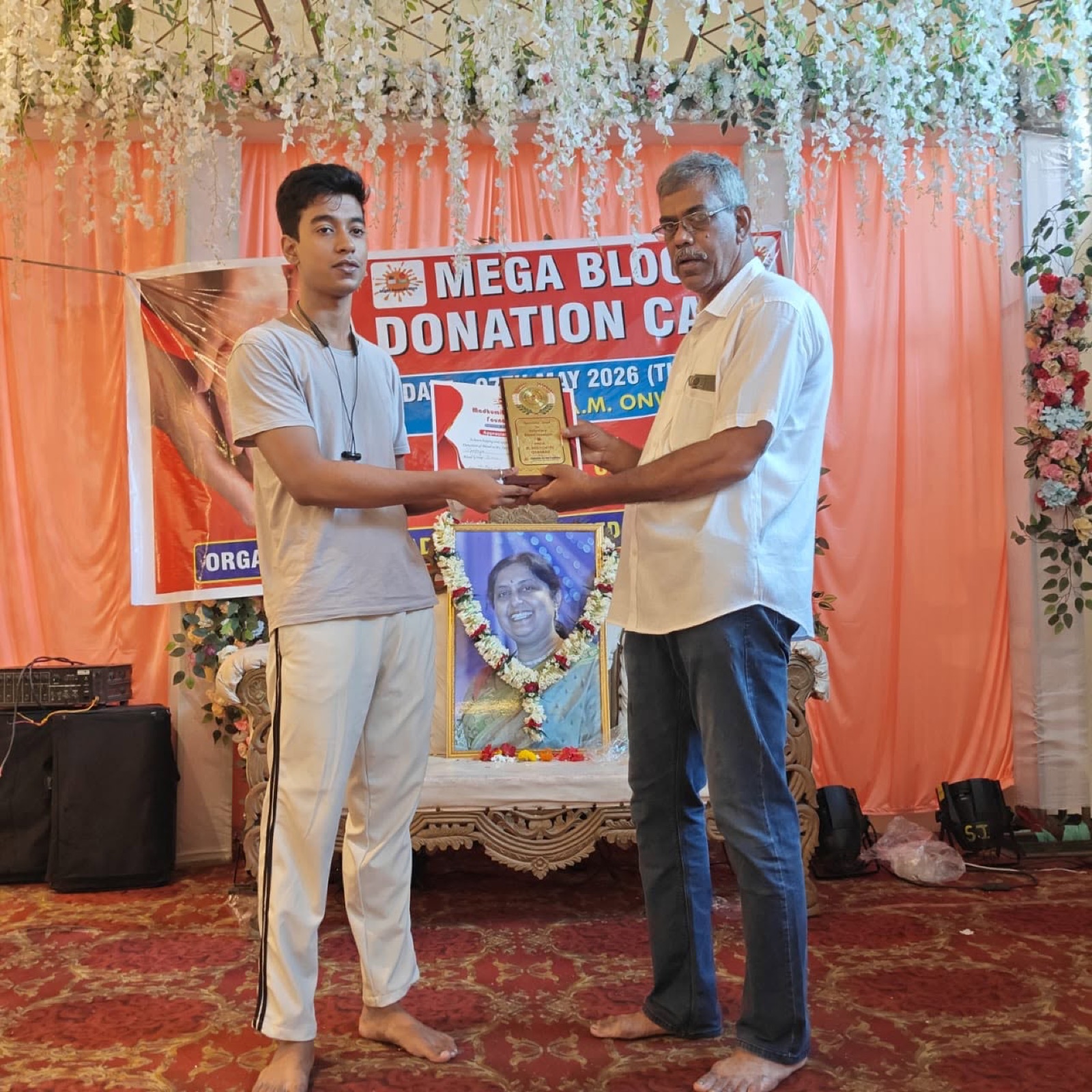 A garlanded portrait of Madhumita honoured on the dais at a camp, with a volunteer receiving a memento